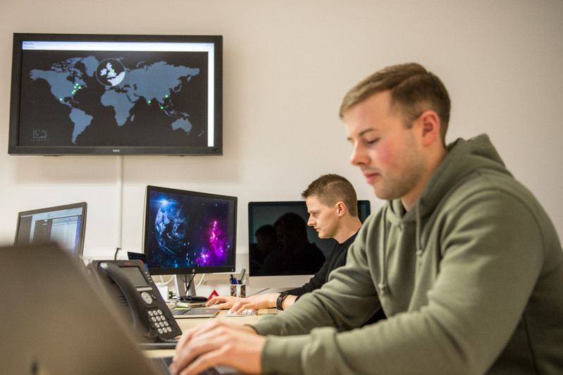 Two men diligently operating computers, situated before a digital display featuring a world map that is illuminated to emphasize specific locations.