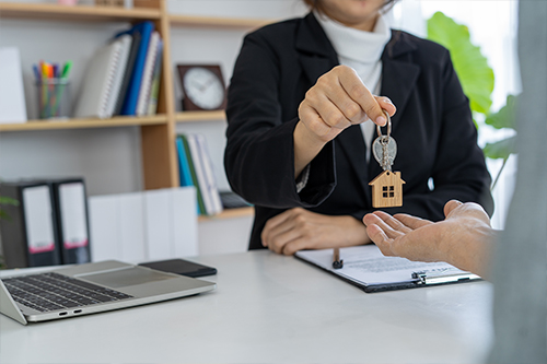 A woman is gracefully passing a set of keys, adorned with a house-shaped keyring, symbolizing the transfer of ownership or rental of a property.