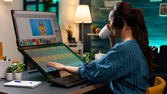 A woman efficiently working within an office setting, sipping on coffee, managing dual screens to access various applications on different devices, all the while enjoying music in the background.
