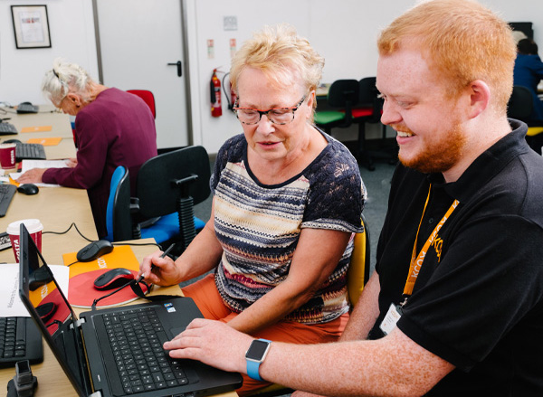 Smiling KCOM employee assists woman in using laptop at an internet drop-in session in library.