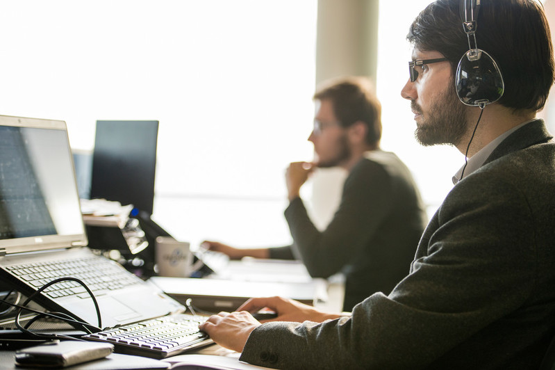 The image features two gentlemen sitting with laptops, with one deeply engrossed in his tasks, while the other is leisurely listening to music, creating a scene of focused work and relaxed enjoyment.